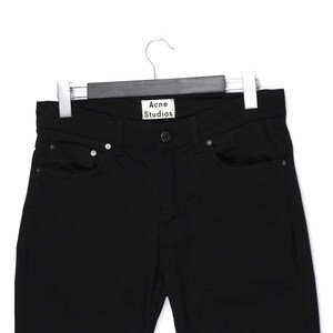 May include: Black denim jeans hanging on a hanger. The jeans have a classic five-pocket design with a button and zipper closure. The brand name "Acne Studios" is visible on a tag at the waistband.