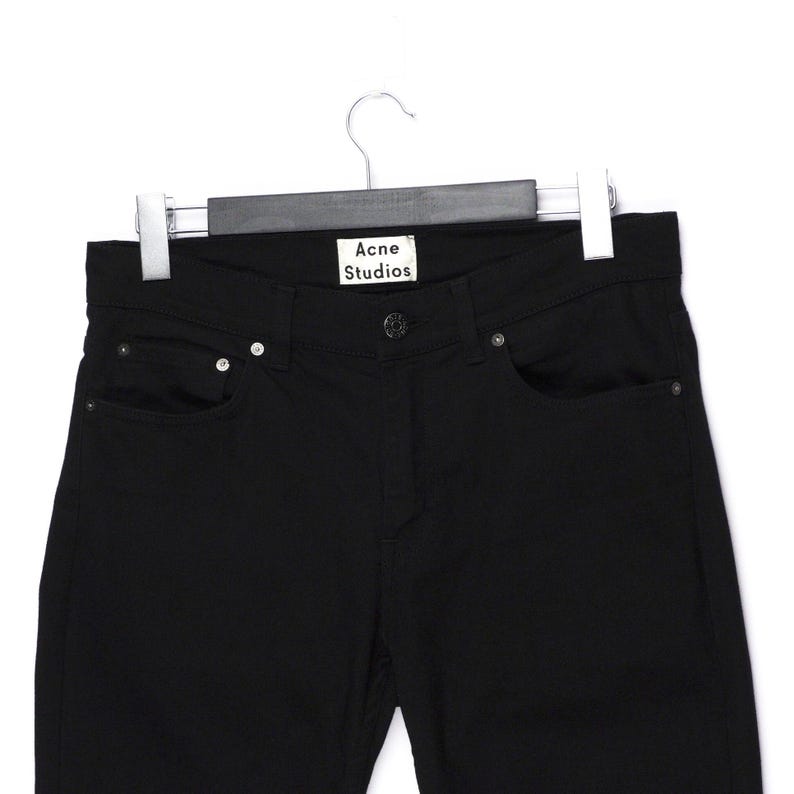 May include: Black denim jeans hanging on a hanger. The jeans have a classic five-pocket design with a button and zipper closure. The brand name "Acne Studios" is visible on a tag at the waistband.