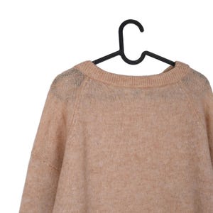Pu&ograve; includere: Un maglione color pesca chiaro con scollo tondo &egrave; appeso a una gruccia di plastica nera. Il maglione ha maniche lunghe e un aspetto morbido e strutturato. Lo sfondo &egrave; bianco, evidenziando il colore e la consistenza del capo.