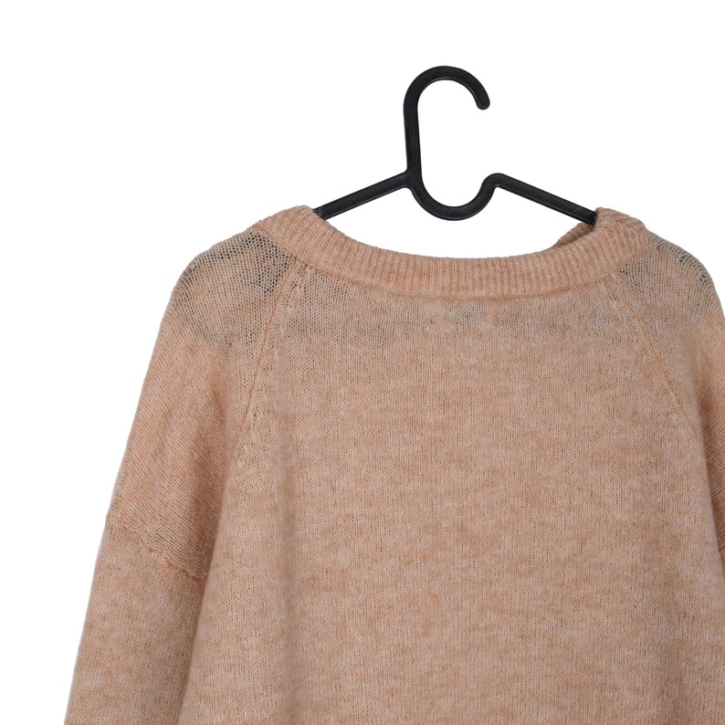 Pu&ograve; includere: Un maglione color pesca chiaro con scollo tondo &egrave; appeso a una gruccia di plastica nera. Il maglione ha maniche lunghe e un aspetto morbido e strutturato. Lo sfondo &egrave; bianco, evidenziando il colore e la consistenza del capo.