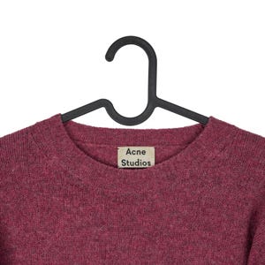 May include: A burgundy crew neck sweater by Acne Studios hangs on a black hanger. The sweater has a textured knit and a small tag with the brand name. The background is plain white.