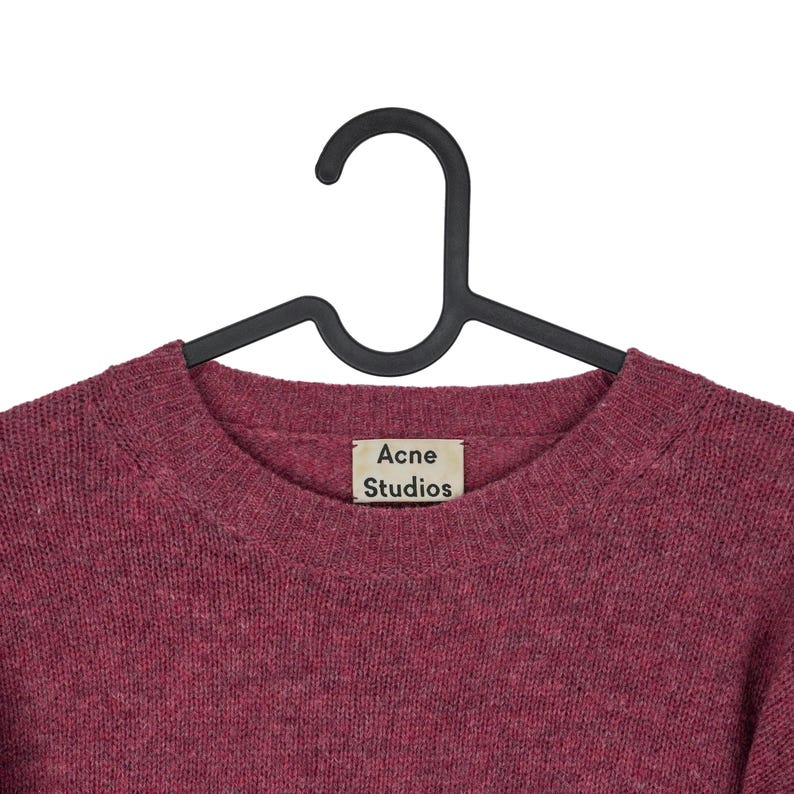 May include: A burgundy crew neck sweater by Acne Studios hangs on a black hanger. The sweater has a textured knit and a small tag with the brand name. The background is plain white.