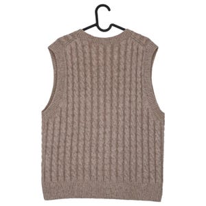 May include: A tan cable-knit sweater vest hangs on a black hanger. The sleeveless garment features a ribbed texture and a rounded neckline. The neutral color and classic design make it a versatile layering piece.