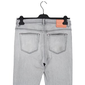 May include: Light gray denim jeans with a peach-colored leather patch on the back, hanging on a black hanger. The jeans have two back pockets and are made of a light-wash denim material.