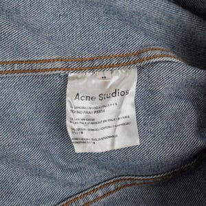 Op de afbeelding: Close-up van een label van een spijkerjasje. Het label luidt "Acne Studios" met extra tekst waaronder "TOP IND FRAY PSS16" en "MADE IN ITALY". De jas is blauw met zichtbare stiksels.