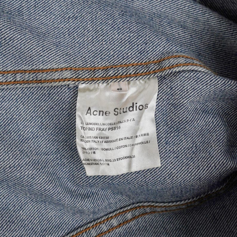 Op de afbeelding: Close-up van een label van een spijkerjasje. Het label luidt "Acne Studios" met extra tekst waaronder "TOP IND FRAY PSS16" en "MADE IN ITALY". De jas is blauw met zichtbare stiksels.