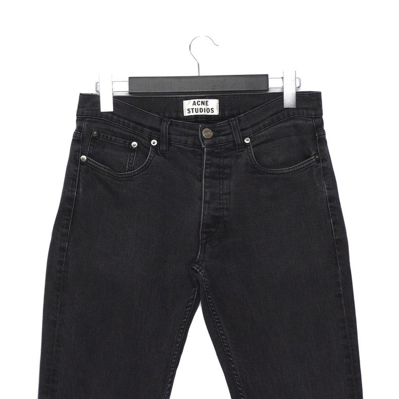 Pu&ograve; includere: Jeans di denim neri appesi a un'appendiabiti. I jeans hanno una chiusura con bottoni, cinque tasche e l'etichetta "ACNE STUDIOS" &egrave; visibile in vita. I jeans sono di un lavaggio scuro e sembrano nuovi.
