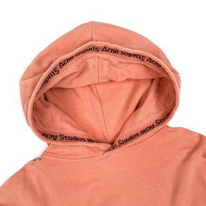 May include: A close-up of a peach-colored hoodie with a drawstring hood. The inside of the hood features the text "Acne Studios" in black lettering. The hoodie is made of a soft material and is set against a white background.