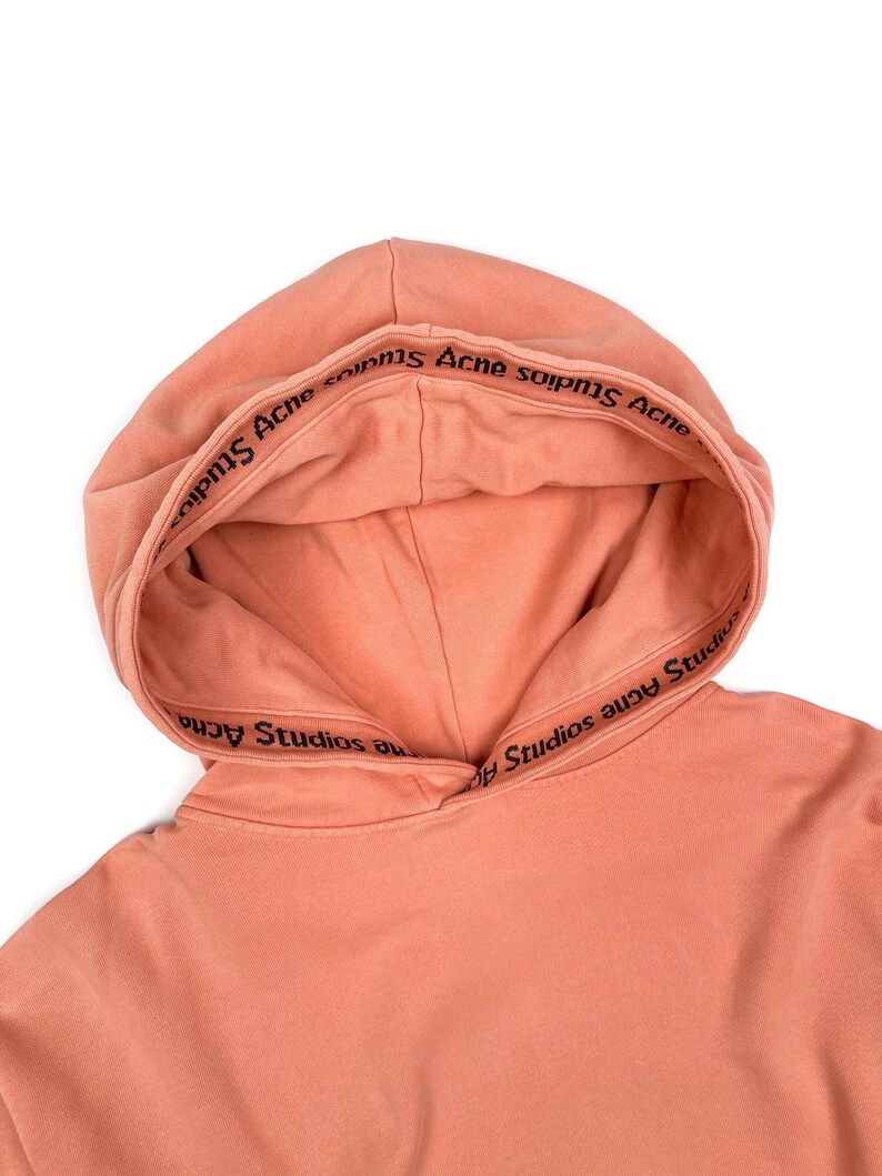 May include: A close-up of a peach-colored hoodie with a drawstring hood. The inside of the hood features the text "Acne Studios" in black lettering. The hoodie is made of a soft material and is set against a white background.
