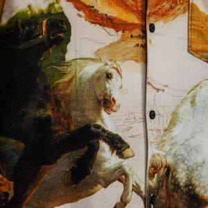 May include: Button-up shirt featuring a detailed print of a classical painting with horses. The shirt has a beige background with black buttons and a pocket. The artwork includes shades of white, black, brown, and orange.