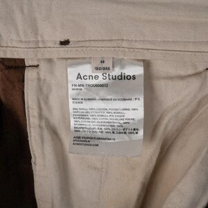 May include: Close-up of a garment tag from Acne Studios. The tag is white with black text, including the brand name, size, and fabric composition. The tag is inside a pair of brown pants.