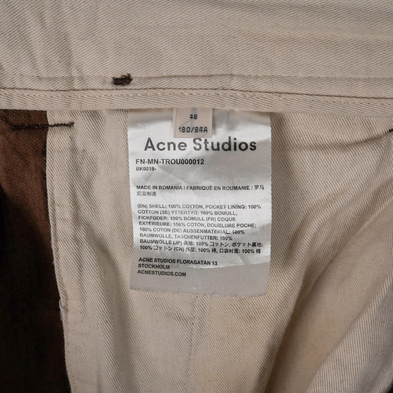 May include: Close-up of a garment tag from Acne Studios. The tag is white with black text, including the brand name, size, and fabric composition. The tag is inside a pair of brown pants.
