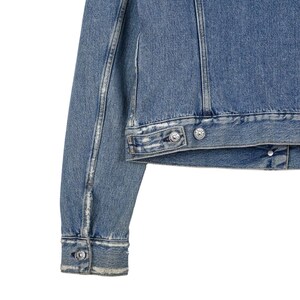 Op de afbeelding: Close-up van een lichtblauwe spijkerjas. De jas heeft zilverkleurige knopen en een klassiek ontwerp. De mouwen en de body zijn zichtbaar, met distressed details. Een tijdloos kledingstuk.
