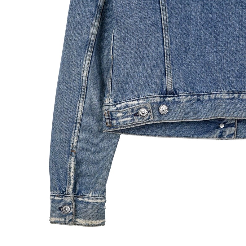 Op de afbeelding: Close-up van een lichtblauwe spijkerjas. De jas heeft zilverkleurige knopen en een klassiek ontwerp. De mouwen en de body zijn zichtbaar, met distressed details. Een tijdloos kledingstuk.
