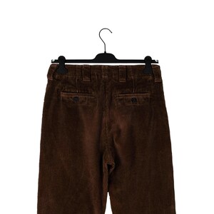 May include: Brown corduroy pants hanging on a black hanger. The pants have two back pockets with button closures and a classic, tailored design. The corduroy fabric has a vertical ribbed texture.