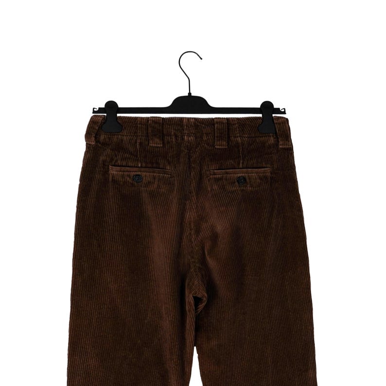May include: Brown corduroy pants hanging on a black hanger. The pants have two back pockets with button closures and a classic, tailored design. The corduroy fabric has a vertical ribbed texture.
