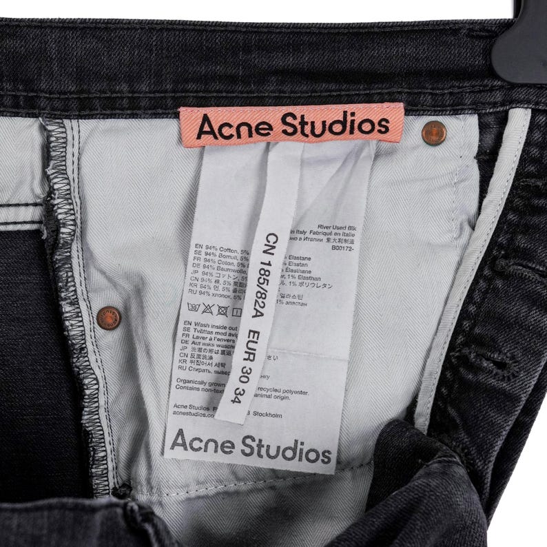 May include: Close-up of the inside of a pair of black denim jeans. The jeans have a light-colored interior and a pink tag with "Acne Studios" printed on it. A white tag with text is also visible, along with copper-colored rivets.