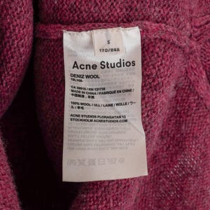 May include: Close-up of a pink wool sweater with a tag from Acne Studios. The tag includes the brand name, size, and material composition. The sweater is made in China.