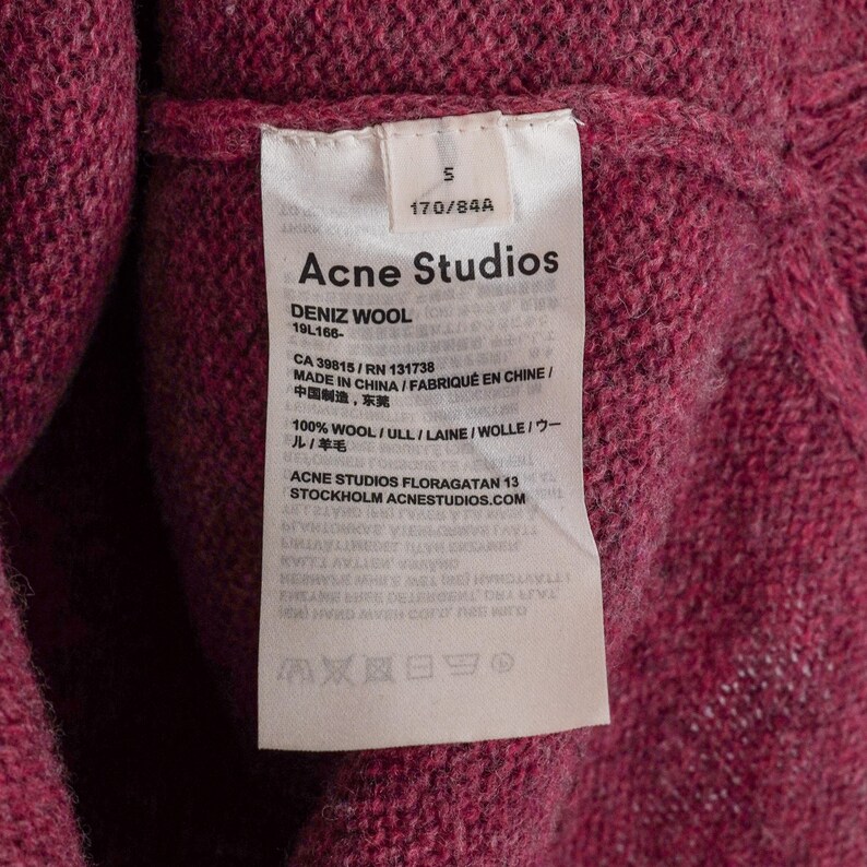 May include: Close-up of a pink wool sweater with a tag from Acne Studios. The tag includes the brand name, size, and material composition. The sweater is made in China.
