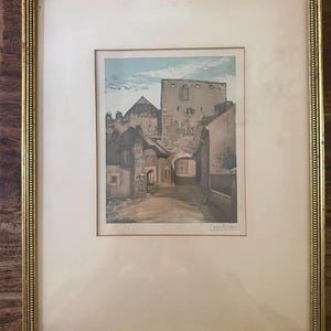 May include: An antique framed print of a European town scene. The artwork depicts buildings and a tower in muted tones of beige, brown, and blue. The print is set within a gold-toned frame and a dark wood outer frame.