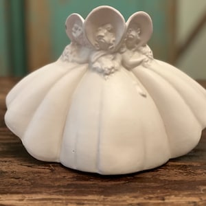 May include: White ceramic figurine of three women wearing hats and dresses. The women are standing together and their dresses are flowing around them.
