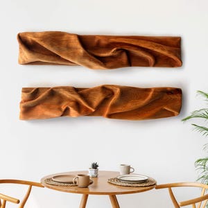 Peut inclure: Deux pièces d'art mural rectangulaires en bois naturel, présentant une texture de surface ondulée et ridée distincte qui imite l'apparence d'un tissu drapé. Le bois affiche un ton brun moyen chaud avec des motifs de grain visibles.