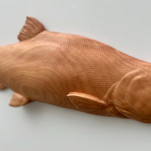 May include: A wooden sculpture of a fish, with a textured surface and fins. The fish is a warm brown colour, with visible scales and a streamlined shape. The artwork is set against a plain white background.