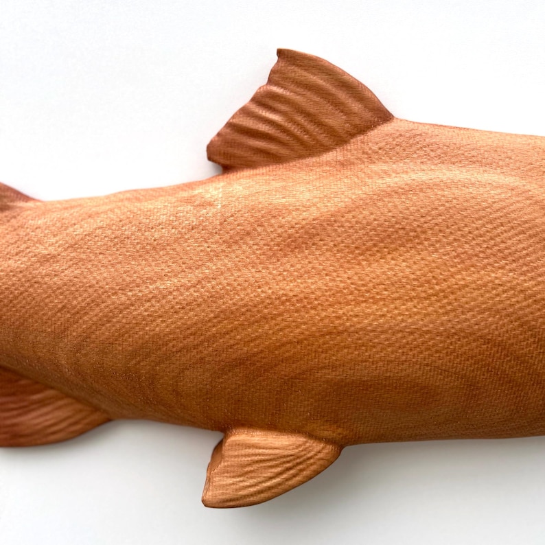 May include: A detailed wooden fish sculpture in a warm reddish-brown hue. The image shows the fish's textured scales and intricately carved fins. The wood grain is visible, adding to the realistic design. The sculpture is a close-up view.