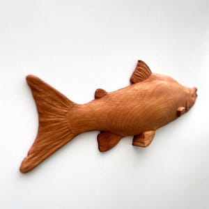 May include: A wooden fish sculpture, intricately carved with a textured surface. The fish is a warm brown hue, with fins, a tail, and a streamlined body. It's likely a decorative piece or wall hanging, crafted from wood.