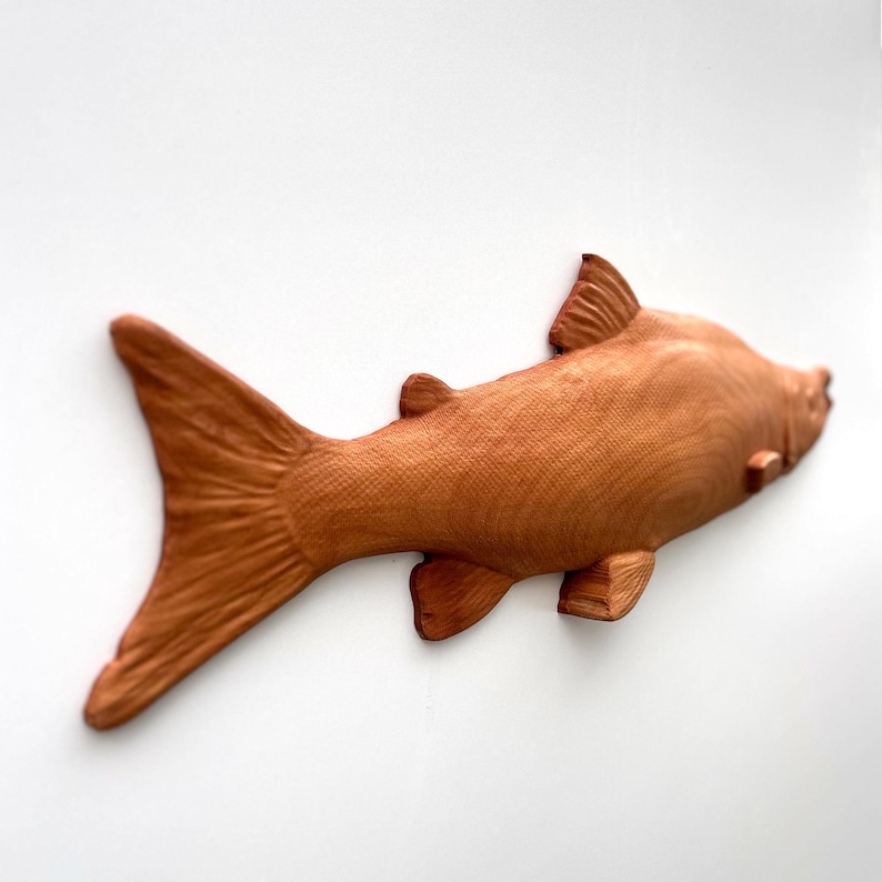 May include: A wooden fish sculpture, intricately carved with a textured surface. The fish is a warm brown hue, with fins, a tail, and a streamlined body. It's likely a decorative piece or wall hanging, crafted from wood.