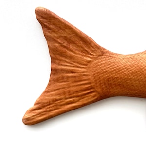 May include: Close-up of a wooden fish tail, showing detailed grain and texture. The tail is a warm, reddish-brown colour, with a fan-like shape. The scales are visible, adding to the realistic appearance. The background is white.