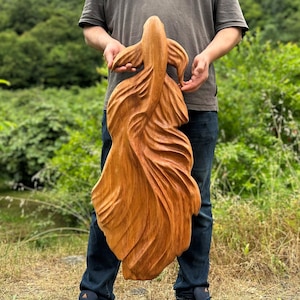 Op de afbeelding: Een grote, handgesneden houten sculptuur van een koikarper. De sculptuur heeft een warme, natuurlijke houttint met gedetailleerd snijwerk van de vinnen en het lichaam van de vis. De koikarper wordt door een persoon voor een groene achtergrond vastgehouden.