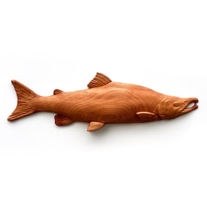 May include: A carved wooden fish sculpture in a natural wood tone. The fish is detailed with fins, a tail, and an open mouth, showcasing the texture of the wood grain. The sculpture is likely for home decor.