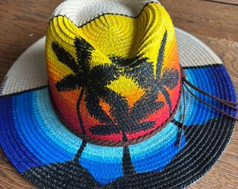 May include: A woven straw fedora hat featuring a hand-painted sunset scene with yellow, orange, and red hues on the crown and a blue ocean gradient on the brim. Black silhouettes of palm trees are painted across the hat, accented by a brown cord.