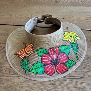 May include: A tan woven straw visor featuring embroidered floral designs. The visor displays a large red hibiscus flower with green leaves, a smaller orange lily-like flower with green leaves, and a yellow flower with green leaves on the brim.