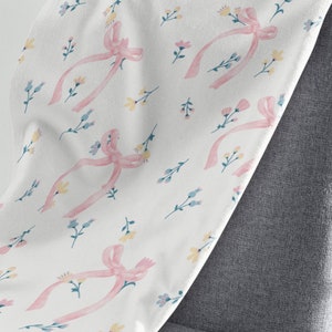 Puede incluir: Una manta blanca con un patr&oacute;n de cinta rosa y flores. La manta est&aacute; colocada sobre una silla gris.
