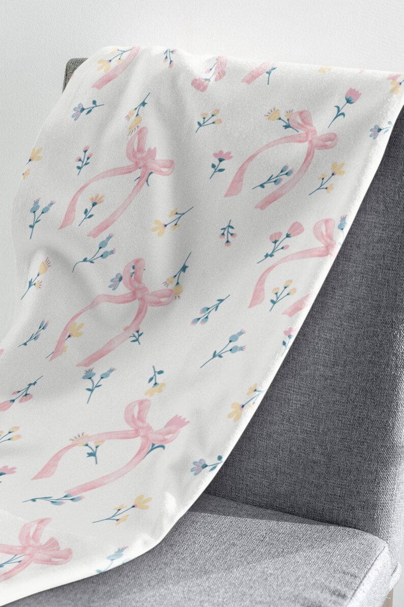 Puede incluir: Una manta blanca con un patr&oacute;n de cinta rosa y flores. La manta est&aacute; colocada sobre una silla gris.