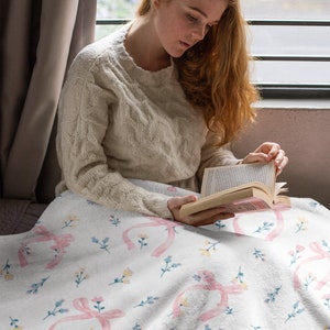 Puede incluir: Una mujer sentada en una cama con una manta blanca con un lazo rosa y un patr&oacute;n floral. Est&aacute; leyendo un libro.