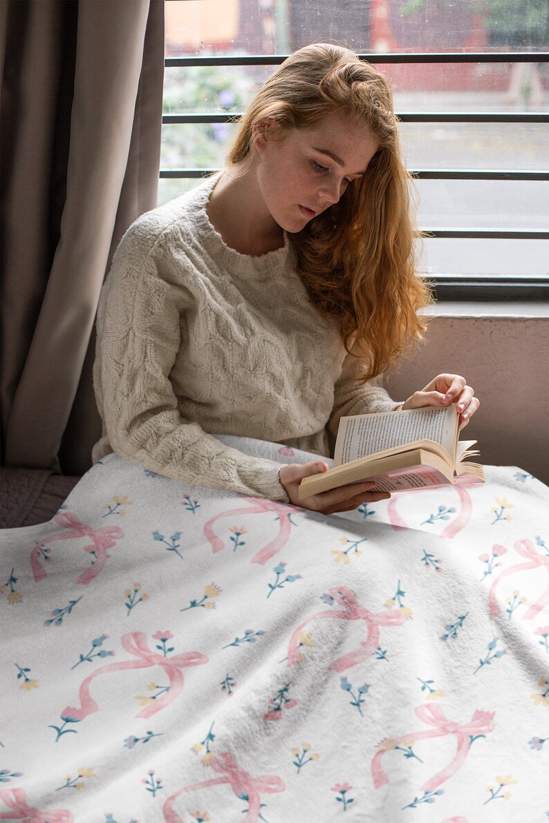 Puede incluir: Una mujer sentada en una cama con una manta blanca con un lazo rosa y un patr&oacute;n floral. Est&aacute; leyendo un libro.