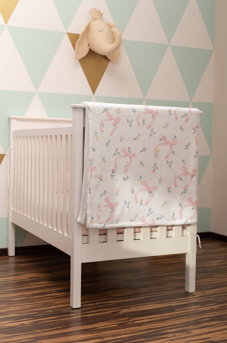 Puede incluir: Cuna blanca con una manta de estampado floral rosa y blanco drapeada sobre el lado. La cuna est&aacute; en una habitaci&oacute;n con una pared de estampado geom&eacute;trico verde claro y blanco.