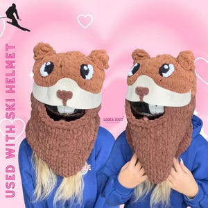 May include: Two brown knitted ski helmet covers in the shape of a beaver with white accents and large teeth. The covers have a brown beard and are worn over a blue hoodie. Text on the left says "USED WITH SKI HELMET".