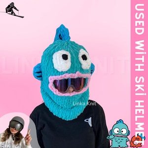 May include: A turquoise blue knitted ski mask with a cartoon fish face. The mask has white eyes, black pupils, a pink mouth, and a black ski goggle. The text "Linka Knit" is visible on the mask.