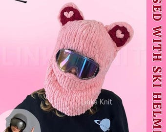 Op de afbeelding: Een roze gebreide skihelmhoes met oorvormige accenten en hartdetails. De hoes heeft een ingebouwde gezichtsbescherming en is ontworpen om te worden gedragen met een skibril. De tekst "Linka Knit" is zichtbaar.