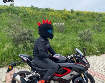 May include: A black motorcycle helmet cover featuring three bright red, upright, triangular spikes on top. The cover is made of a textured knit material and fits over a full-face helmet with a blue tinted visor. The text LINKA KNIT is visible.