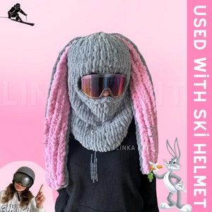 May include: Gray knitted ski mask with pink bunny ears. The mask has a built-in face covering and eye holes. The text "USED WITH SKI HELMET" is visible in the image.