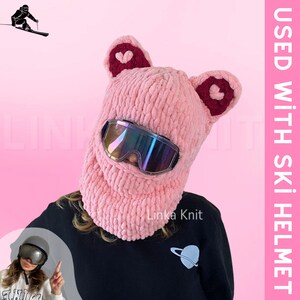 May include: Pink knitted ski mask with ear flaps and heart details. The mask is worn over a person's face and covers the nose and mouth. The person is wearing ski goggles.