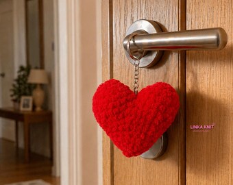 May include: A plush, red heart-shaped keychain hanging from a silver door handle. The heart is made of a soft, textured material. The door is made of wood.