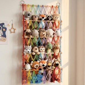 May include: A wall-mounted storage organiser featuring a rainbow-coloured macrame net suspended from wooden dowels. The net holds a collection of various plush animal toys, including a koala, fox, owl, and dinosaur, arranged across five horizontal tiers.