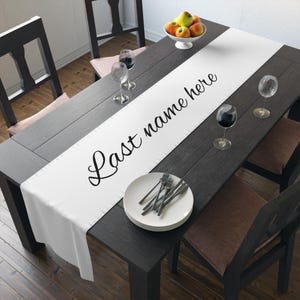 May include: A white table runner featuring the black cursive text "Last name here" centred along its length. The runner is laid out on a dark wood dining table, which is set with white plates, silverware, wine glasses, and a bowl of apples.