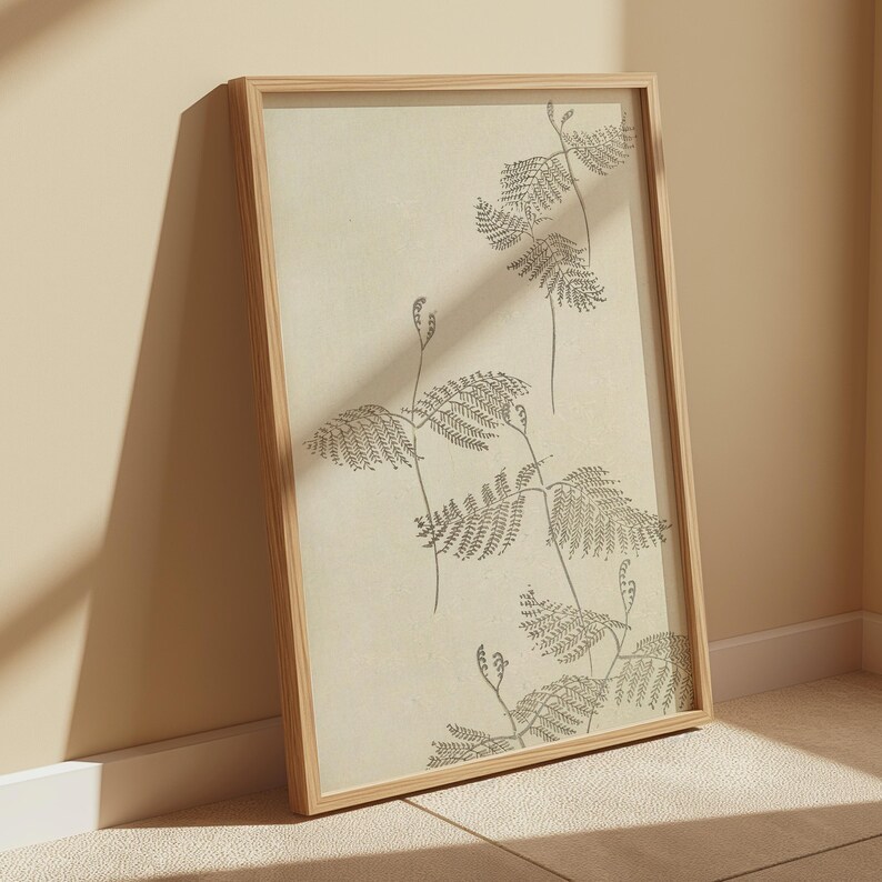 May include: A framed botanical print featuring a detailed illustration of fern-like plants in shades of gray against a cream-colored background. The artwork is encased in a natural wood frame, leaning against a wall. The overall aesthetic is minimalist and nature-inspired.